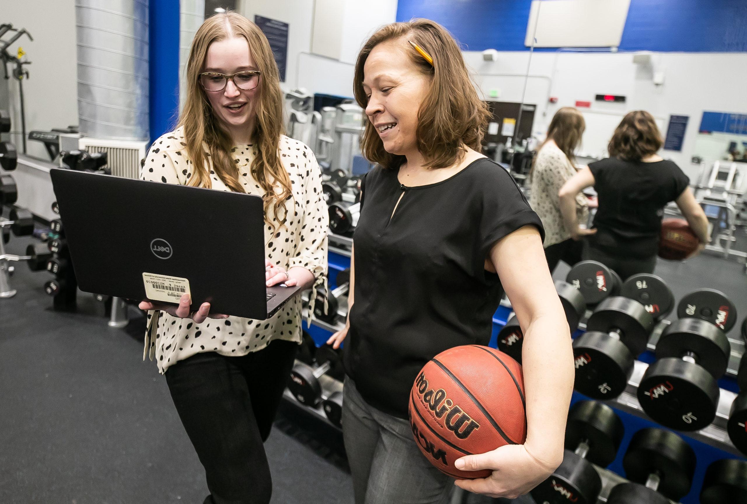 People standing in a gym, one is holding a basketball and the other a laptop