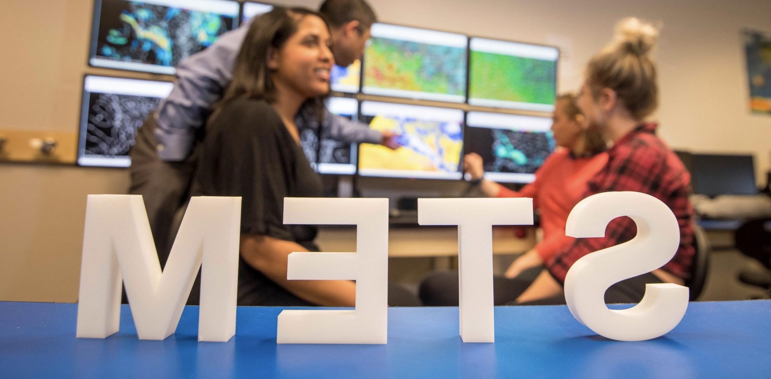 STEM sign on a blue table with three female students and a male instructor behind it discussing weather on computer displays