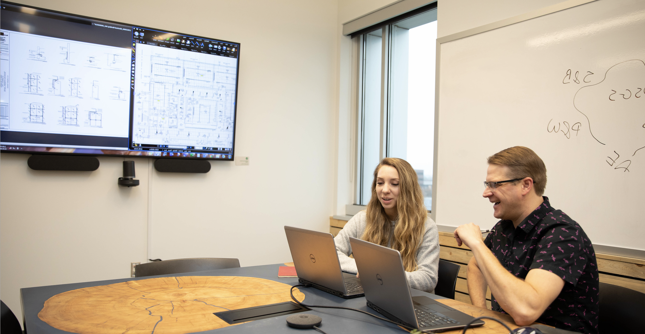 Two people sitting in a conference room at laptops with screens behind