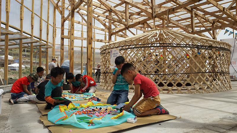 A group of children sit on the floor of a wood-framed and glass-clad structure. The children are interacting with colorful materials.