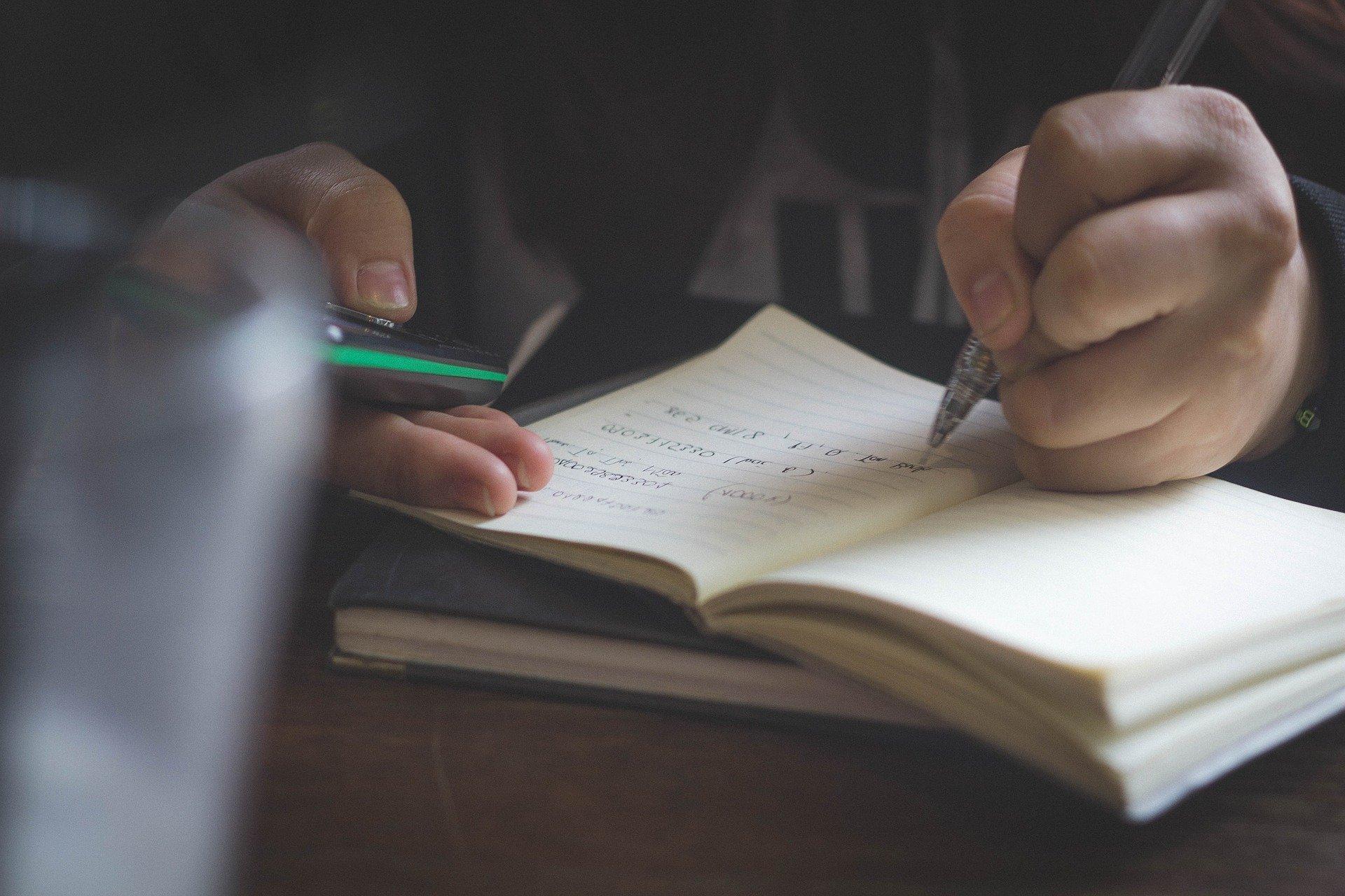 A person writing in a notebook and holding a phone