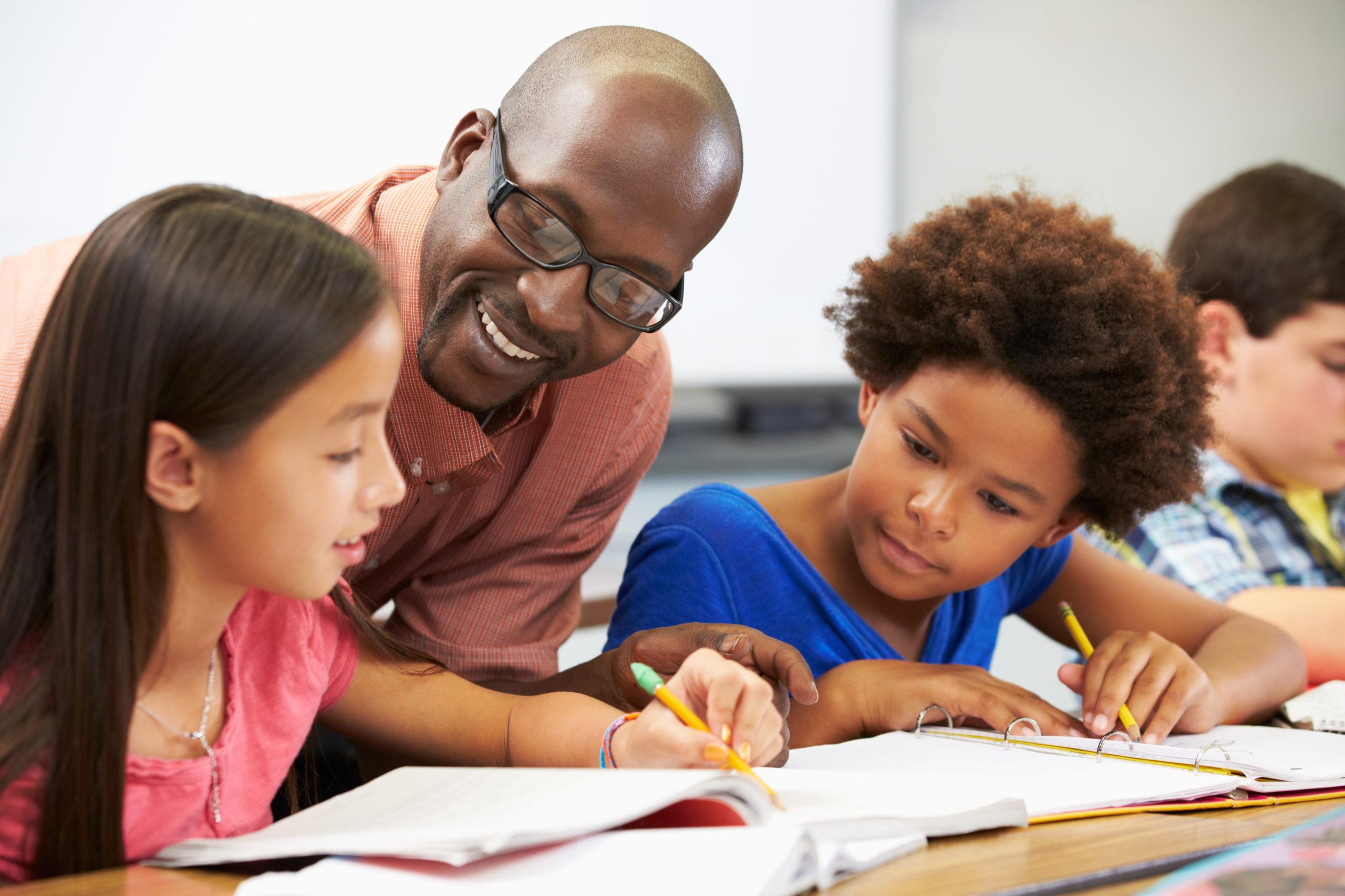 Teacher helping students studying at desks In classroom checking work and smiling.