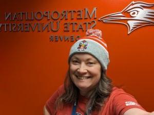 Monica woman with brown hair in a Rowdy hat standing in front of a red wall with metallic MSU Denver logo