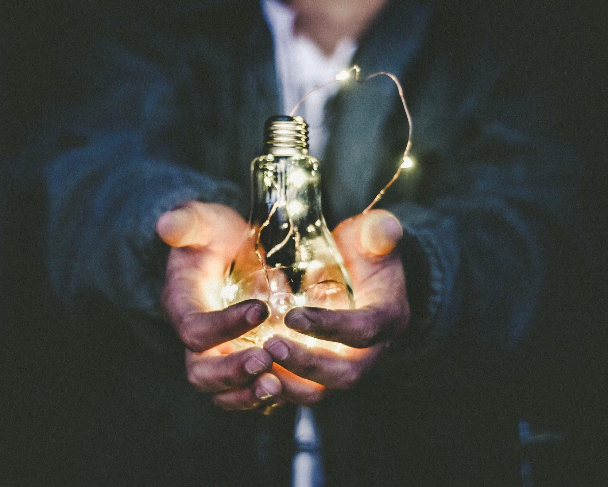 A pair of hands holding a light bulb with a string of lights illuminating the bulb