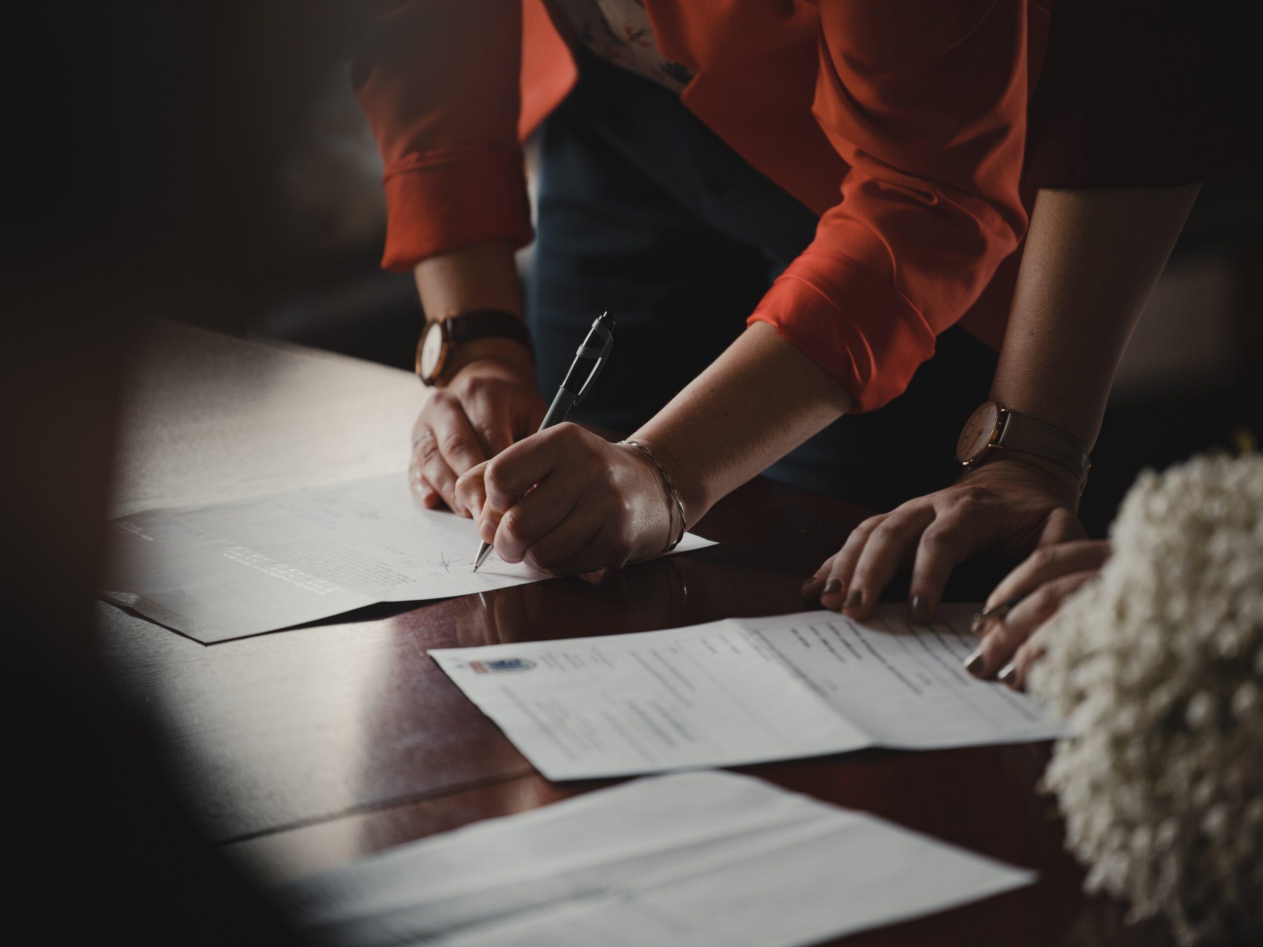 Two sets of hands with pieces of paper on a table, one person is signing one of the pieces of paper