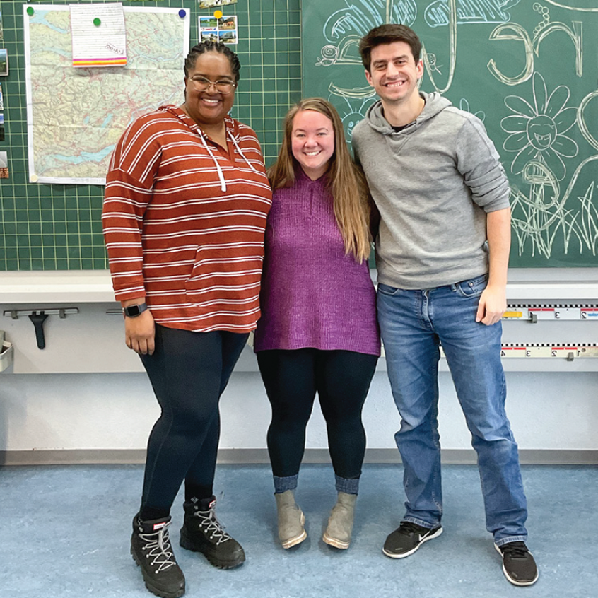 Three students smiling, standing together in front of a chalkboard in a classroom.