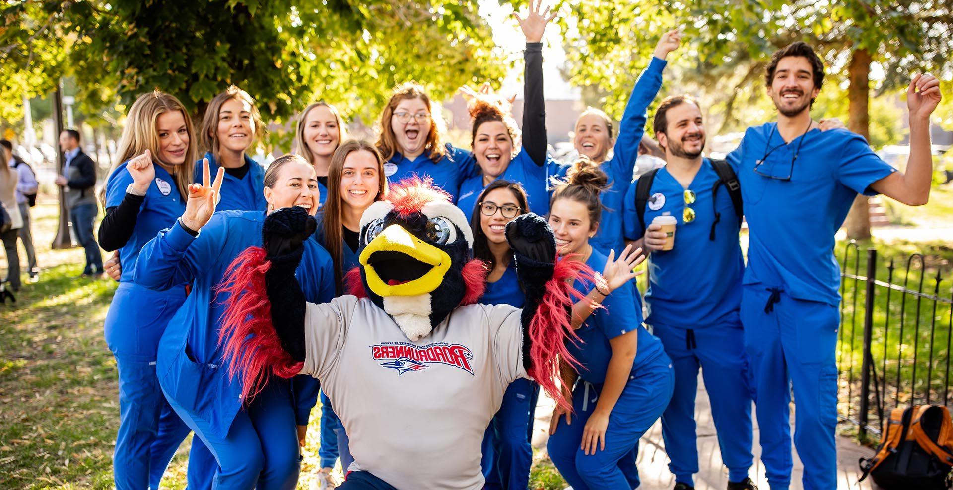 Healthcare students in royal blue scrubs posing for a picture with Rowdy on a sunny, fall day