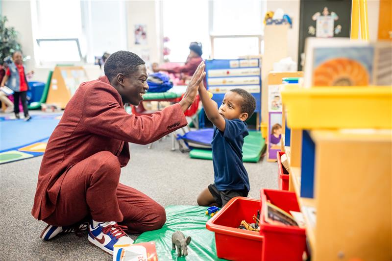 A photo of a MiSTER kneeling on the floor of a classroom, high fiving a young student.