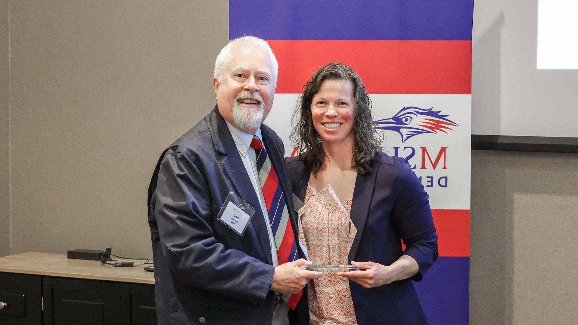 Paul Larson ('72) standing with Brandi Rideout and holding a glass award at the 2023 Golden Nest Club luncheon