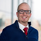 Headshot of Cory Phare, wearing a blue sweater over a white button-up with a red tie and stylish (for 2016) glasses.