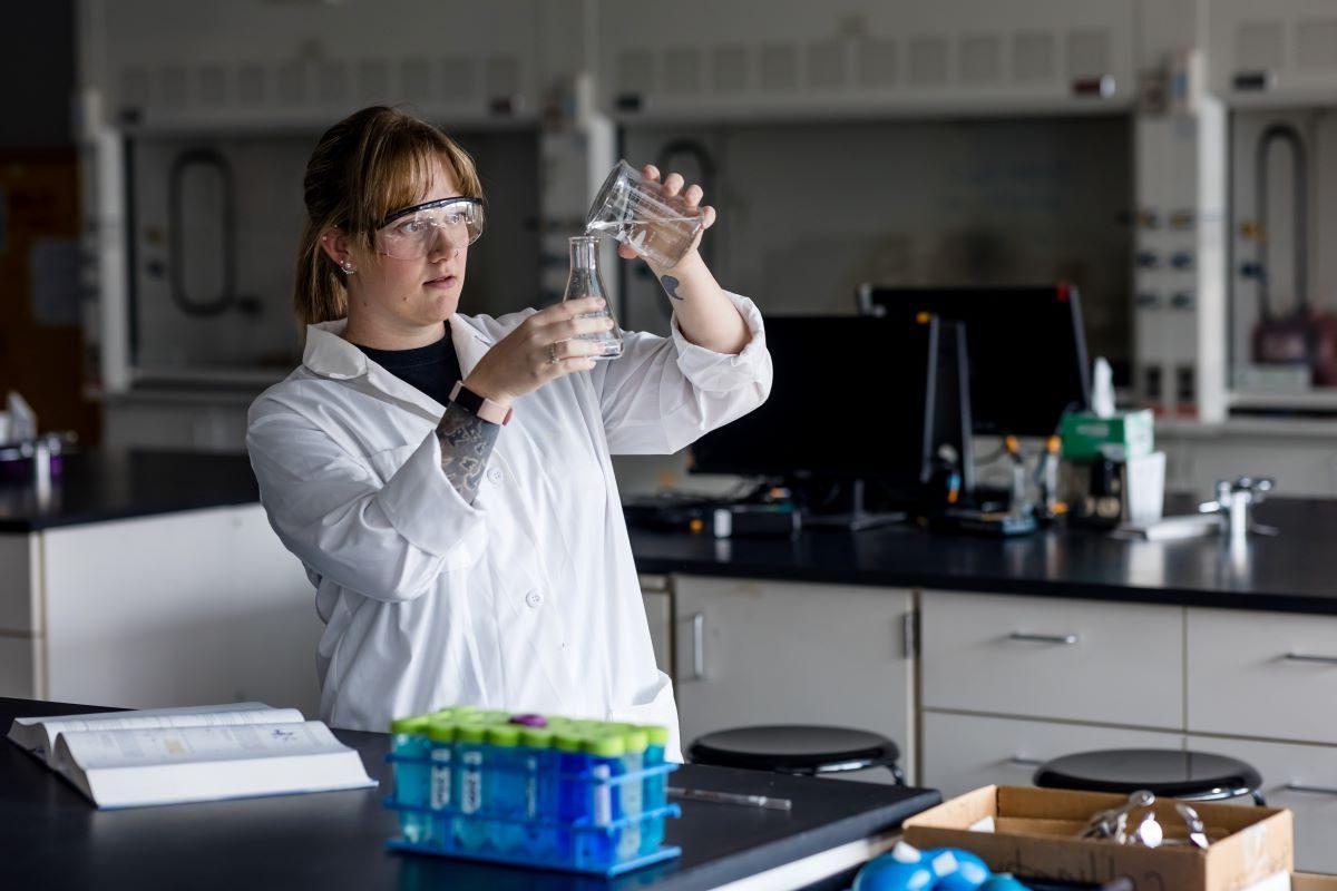 A student wearing a white lab coat and safety goggles pours a clear liquid into a glass beaker in a lab