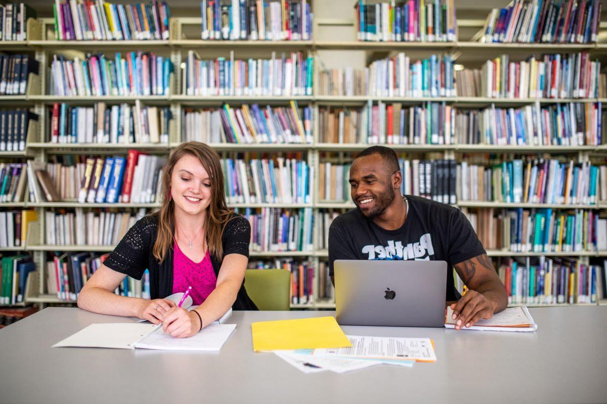 Two students smile as they study in the library with shelves of books behind them