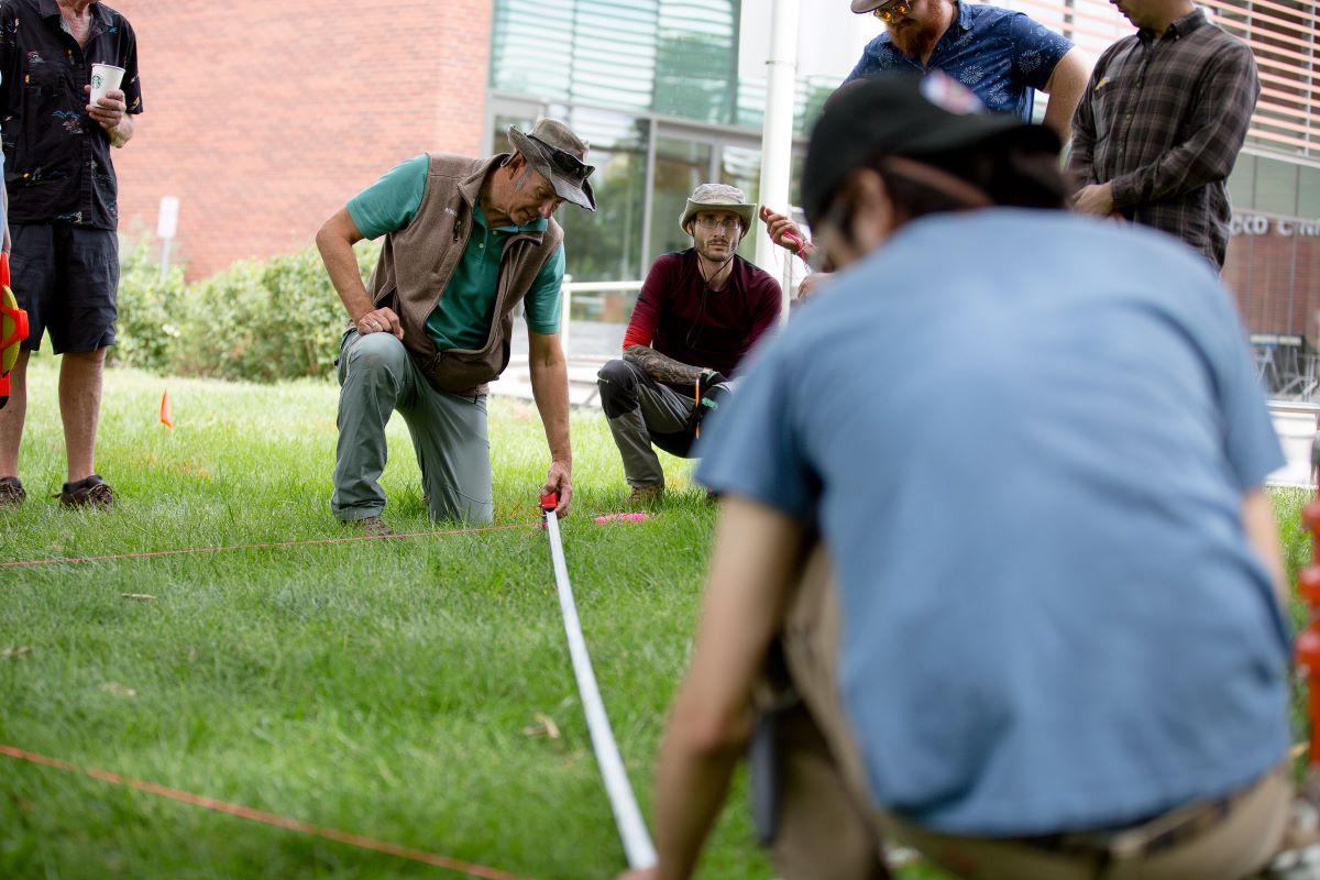 A MSU Denver professor and students kneeling and measuring a grassy area with a tape measure and red string