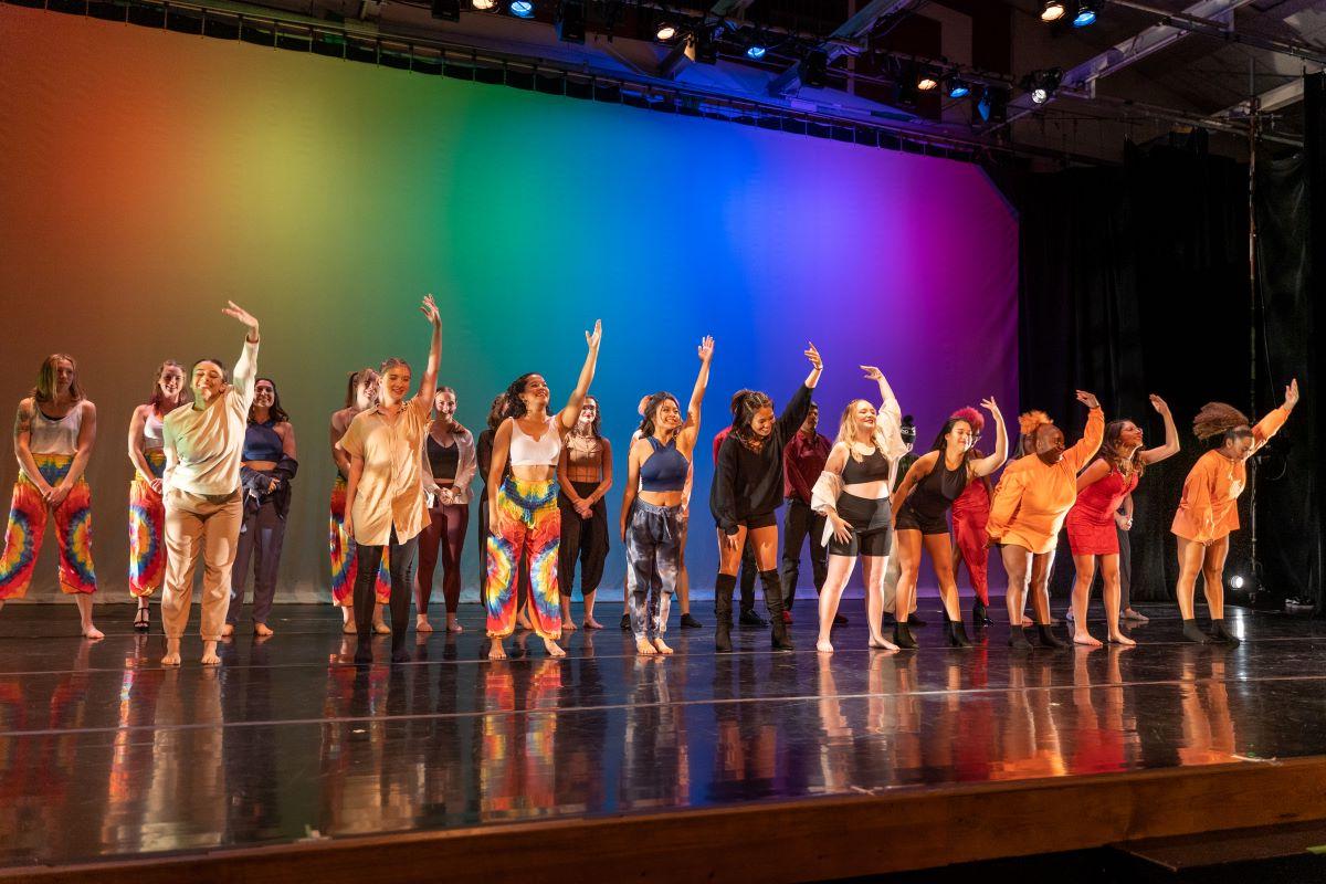 Students in costume hold one hand in the air as they bow after a performance in front of a rainbow backdrop